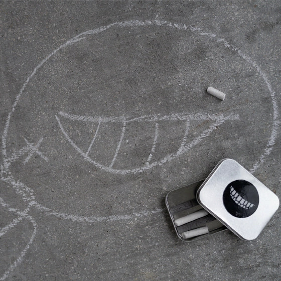 White smiling face symbol written in chalk on gray pavement with an open silver tin of chalk next to it