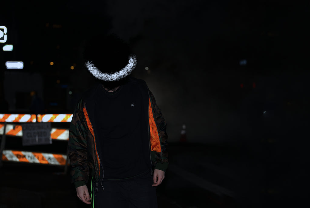 Person walking on a street at night with a green hanging thread visible and face covered