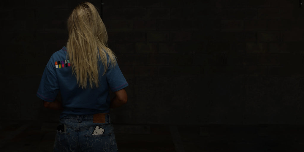 A woman with blonde hair facing away from the camera wearing a blue normal;c t-shirt with colorful patches in the shape of pills on the back against a dark background