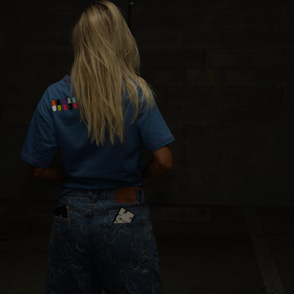 Woman with blonde hair facing away from the camera wearing a blue normal;c shirt with colorful patches in the shape of pills on the back against a dark background.